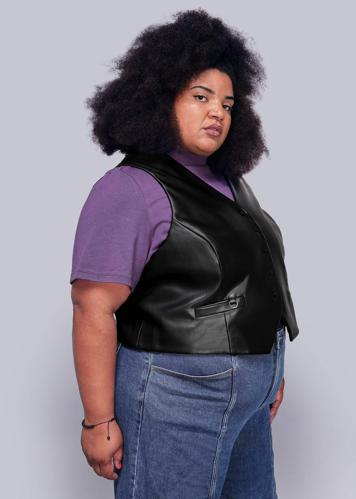 1X=Person wearing a black vegan leather vest over a purple shirt with blue jeans on a light gray background