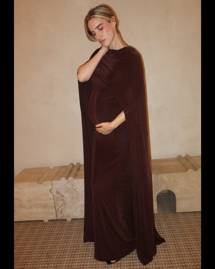A pregnant woman wearing a Bumpsuit maxi dress with a floor length cape detail in chocolate brown against a neutral setting