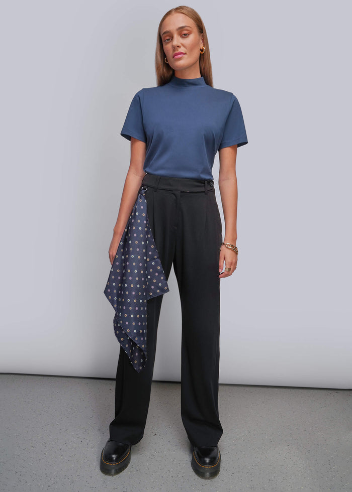 A woman modeling a slate blue mock neck short-sleeve top, black wide-leg trousers, and a navy blue polka dot scarf hanging from their pants, standing against a light gray studio background.