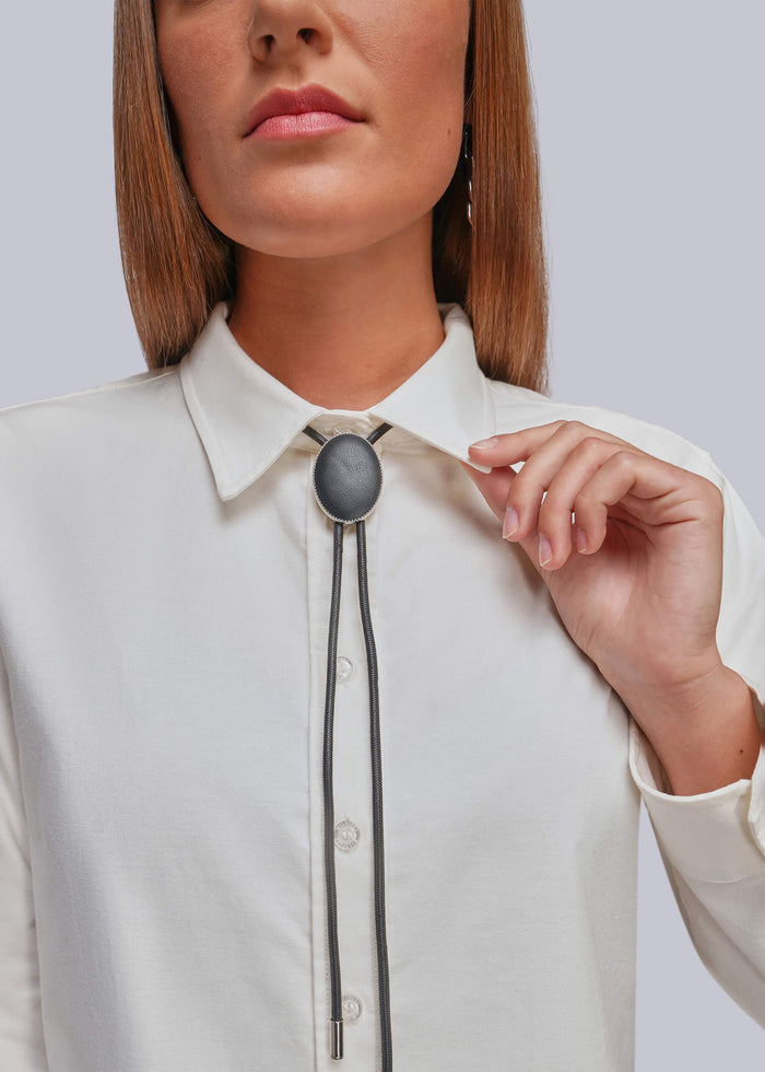 A close-up of a model wearing a white collared button-up shirt and a black bolo tie with a dark gray oval leather centerpiece, set against a solid light gray background.