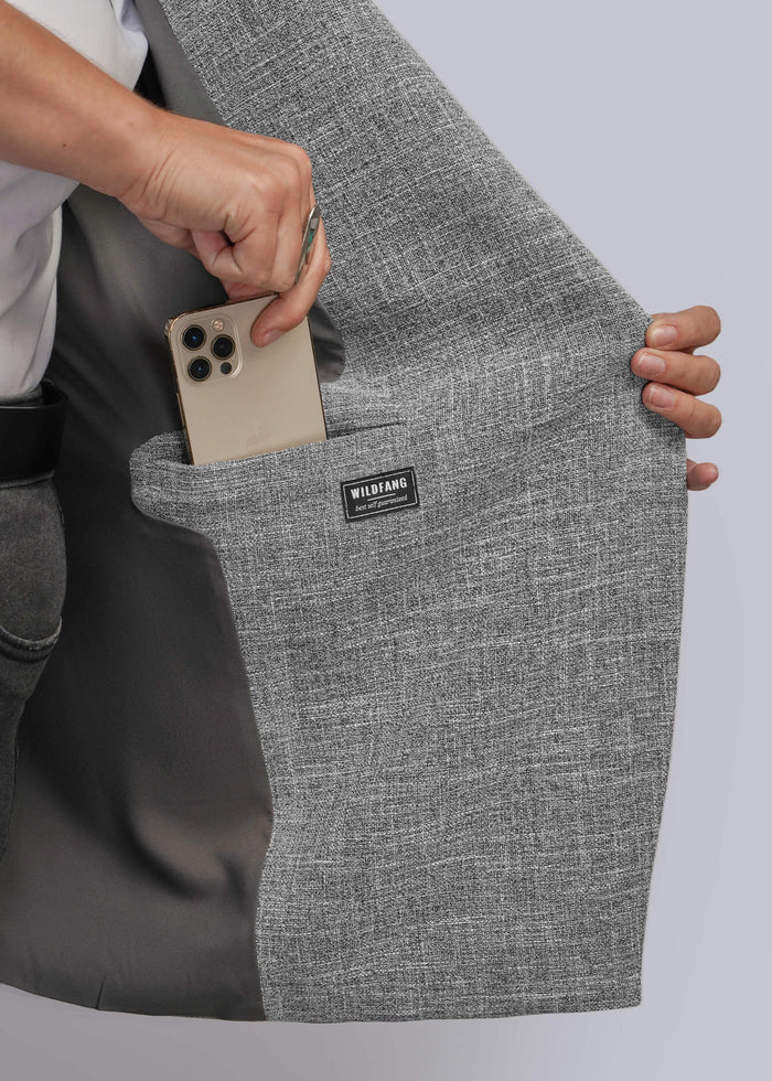 1X=Close-up of a person holding open a gray textured blazer to show a large interior pocket containing a smartphone. A small black brand label reading WILDFANG is visible on the pocket.