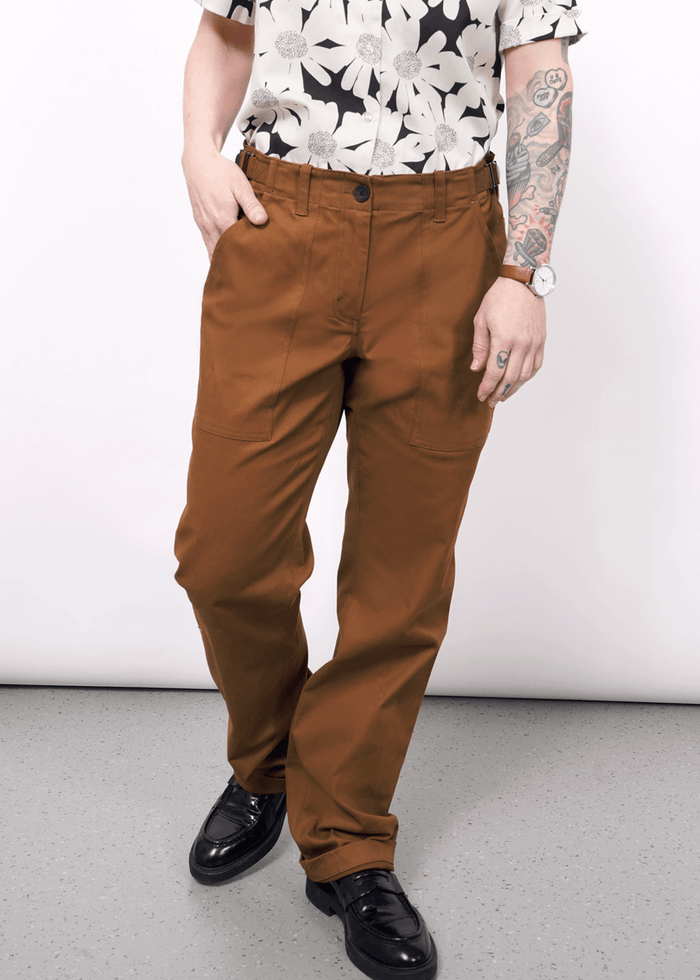 S=A person stands against a light background wearing The Essential Utility Pant in rust, featuring spacious pockets, paired with black loafers and a black-and-white floral shirt. One hand is in a pocket; the other arm with tattoos rests by their side.