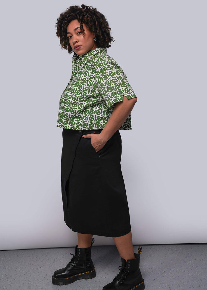 1X=Person wearing a green patterned shirt and black skirt against a white background