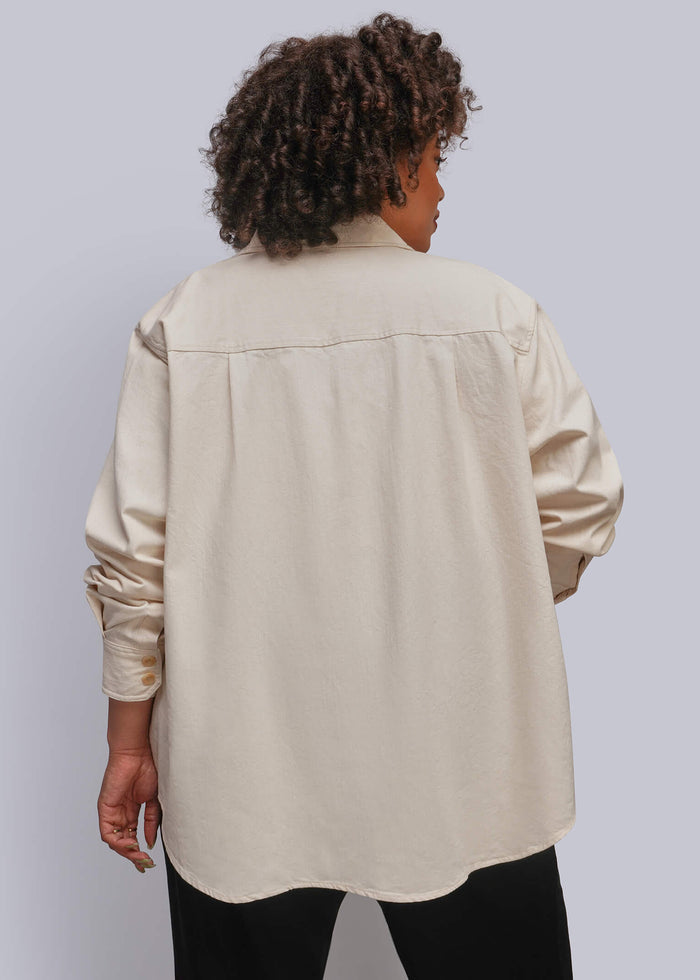 1X=A person shown from the back wearing an oversized cream-colored long-sleeve button-up shirt with black pants, set against a solid light gray studio background.
