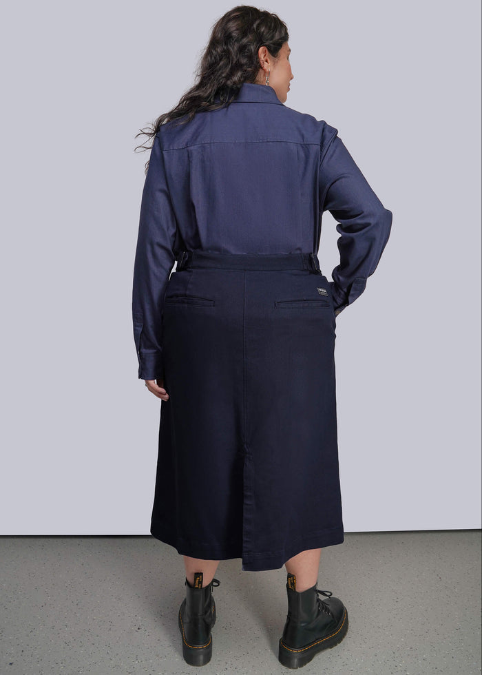 1X=A model is shown from the back wearing a navy blue long-sleeve collared shirt tucked into a matching navy midi skirt, paired with black boots, against a light gray studio background.