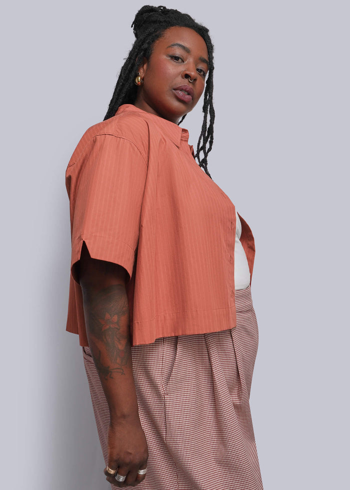 1X=A woman modeling a short-sleeve coral pink cropped top paired with a red and white herringbone pants, shown from a side profile against a solid gray studio background.