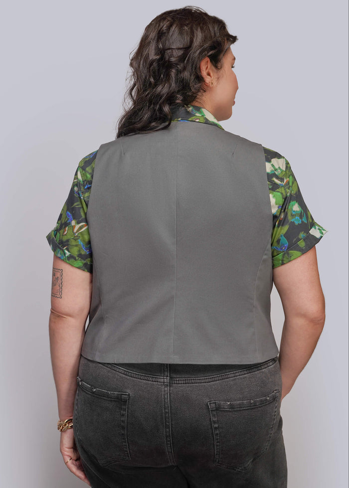 1X=A person shown from the back wearing a sleeveless gray vest over a green and blue floral short-sleeve shirt, paired with dark gray jeans, against a light gray studio background.