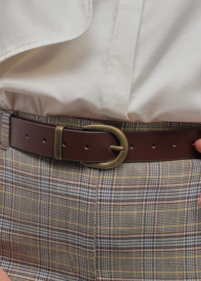 A close-up of a dark brown leather belt with a brass buckle, worn over a cream shirt and a beige and brown plaid skirt.