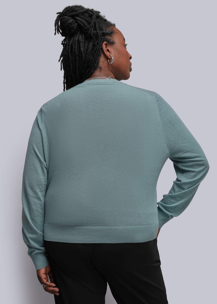 1X=A woman shown from the back wearing a muted sage green long-sleeve knit top and black pants against a plain light gray studio background.