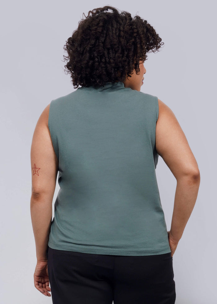 1X=A person with curly hair, wearing The Essential Mock Neck Tank in green, black pants, and black shoes, stands facing away from the camera against a plain white background.