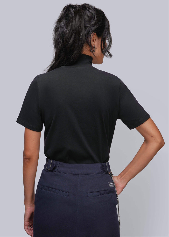 S=A woman shown from the back wearing a black short-sleeve mock neck top and dark navy denim skirt, photographed against a solid light gray studio background.