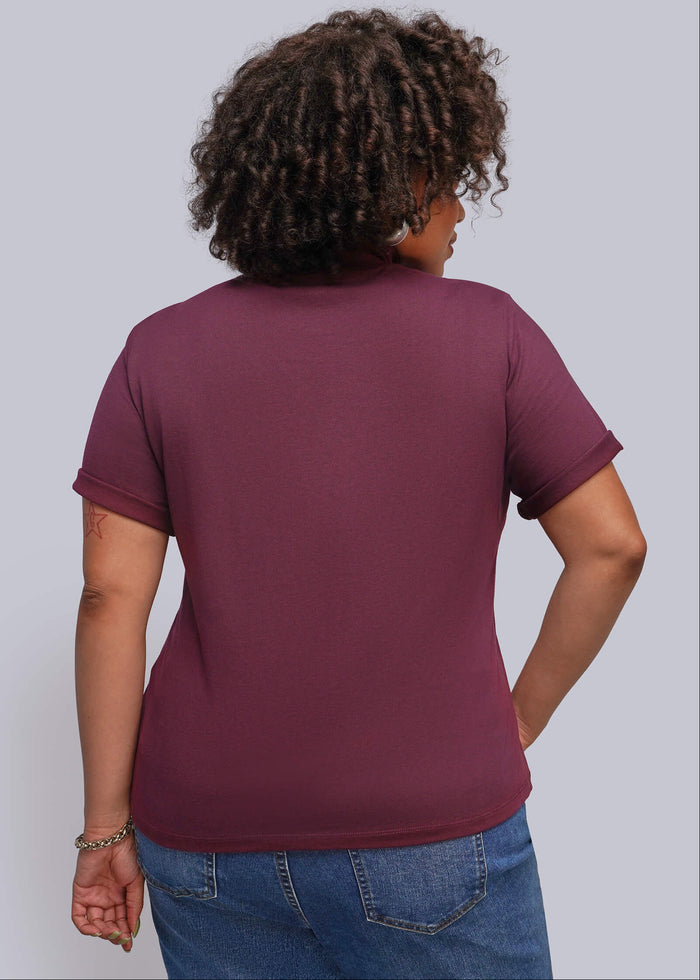 1X=A woman with curly dark hair is shown from the back wearing a short-sleeve t-shirt in a deep burgundy/maroon color, paired with blue jeans, against a solid light gray background.