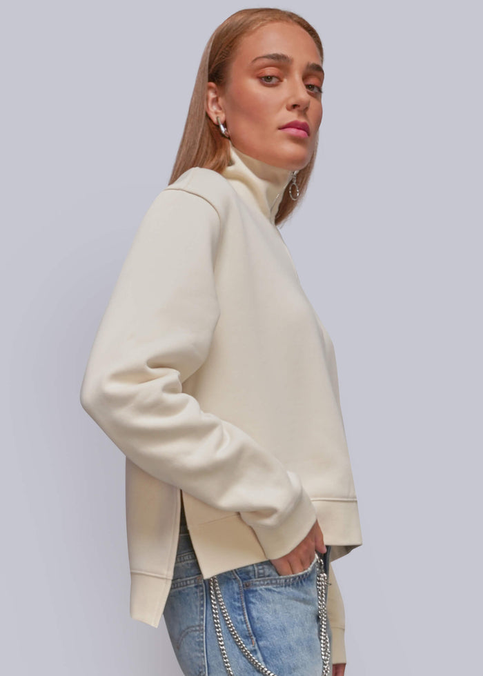 S=A woman wearing a cream-colored, oversized long-sleeve mock-neck sweatshirt and light-wash blue jeans with a silver chain detail on the pocket, posed against a solid light gray studio background.