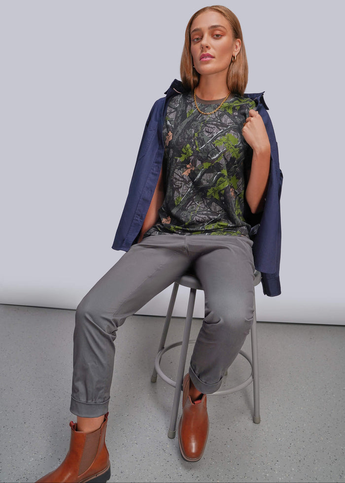 S=A woman seated on a metal stool wearing a dark blue button up draped over a camouflage-patterned top, gray pants, and brown ankle boots against a plain light gray studio background.