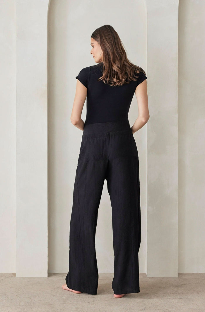 A back view of a pregnant woman wearing white relaxed pants and a black shirt by Bumpsuit against a beige feature wall.