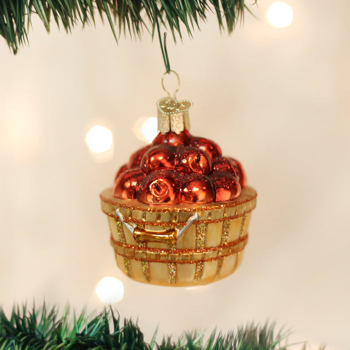 A close-up of a Christmas tree ornament shaped like a basket filled with shiny red apples, hanging on a green tree branch with warm white lights blurred in the background.