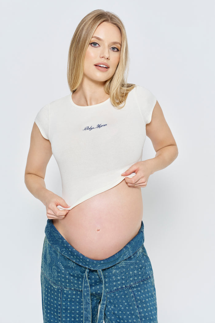 Pregnant woman wearing a white Bumpsuit baby mama tee and blue pants against a light gray background