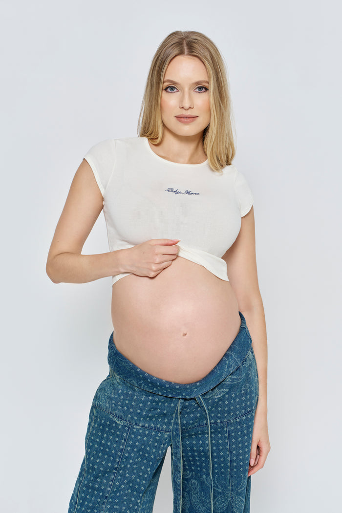 Pregnant woman wearing a white Bumpsuit baby mama tee and blue pants against a light gray background