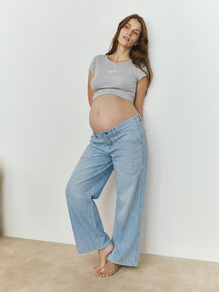 Pregnant woman wearing a Bumpsuit Heather grey baby mama tee and blue jeans against a white wall.