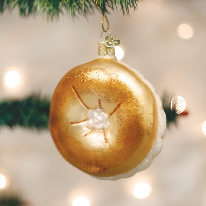 A Christmas tree ornament shaped and painted like a golden brown bagel with cream cheese, hanging from a green branch with blurred holiday lights in the background.