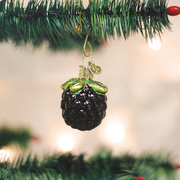 A glossy blackberry-shaped Christmas ornament with green leaves hangs from a gold hook on a green Christmas tree branch, with blurred lights in the background.