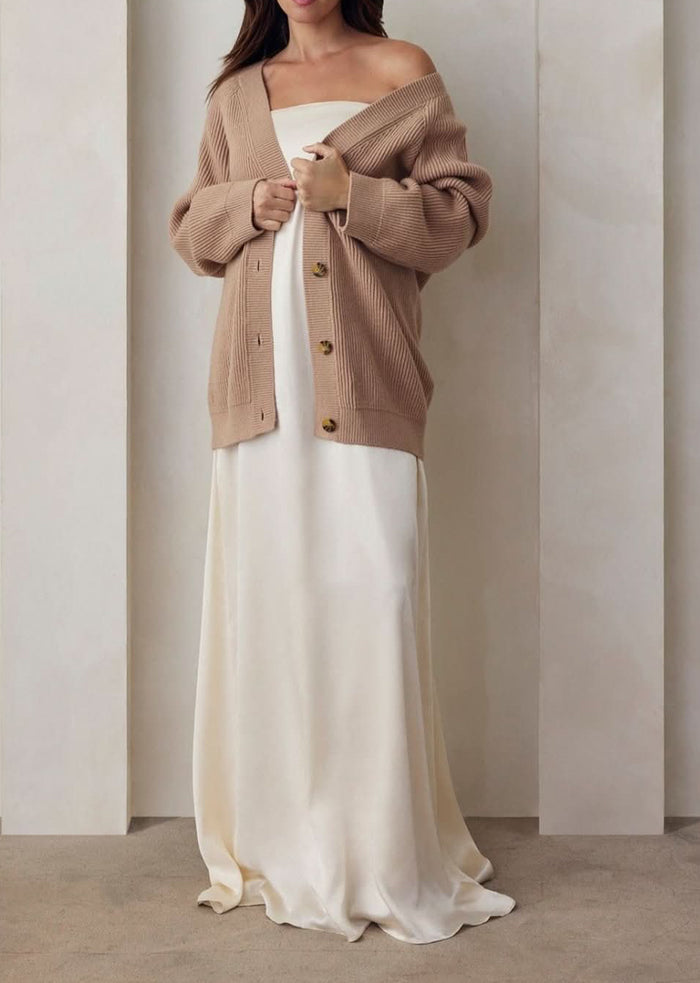 Pregnant model wearing the Bumpsuit brushed satin gown in ivory and a beige knit oversized cardigan against a beige feature wall.