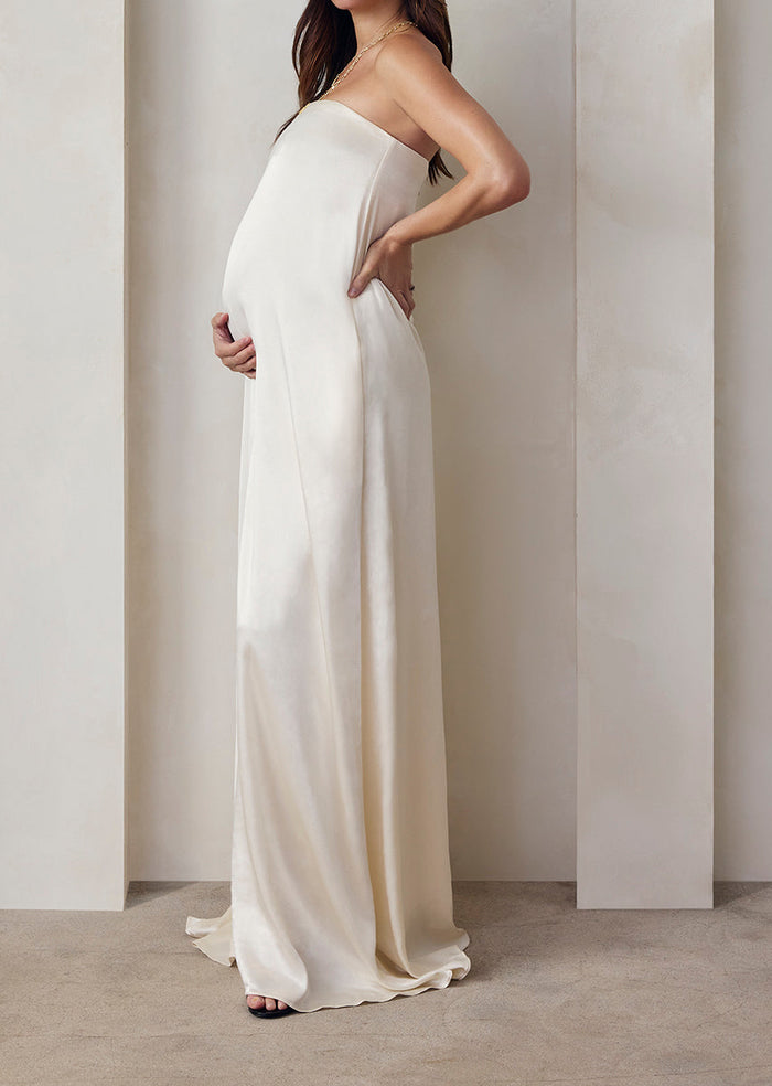 Pregnant model wearing the Bumpsuit brushed satin gown in ivory against a beige feature wall.