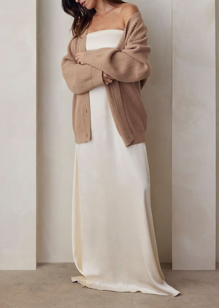 A Pregnant model wearing the Bumpsuit brushed satin gown in ivory and a beige cardigan against a beige feature wall.