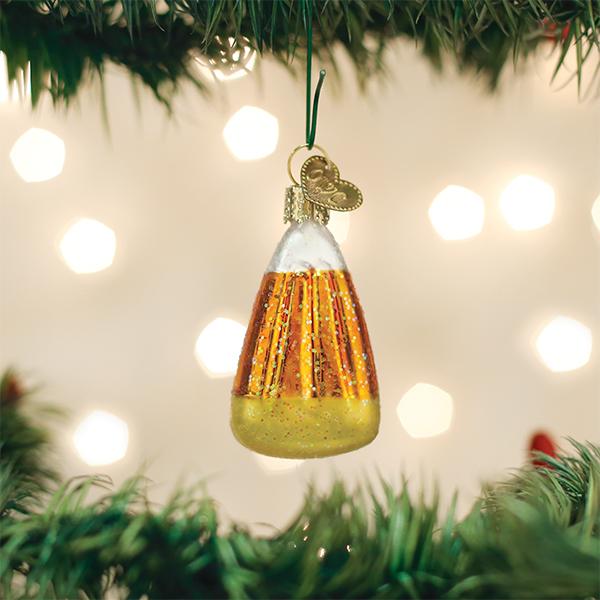 A candy corn-shaped ornament with glittery orange, yellow, and white stripes hangs from a Christmas tree branch, with soft, out-of-focus lights glowing in the background.