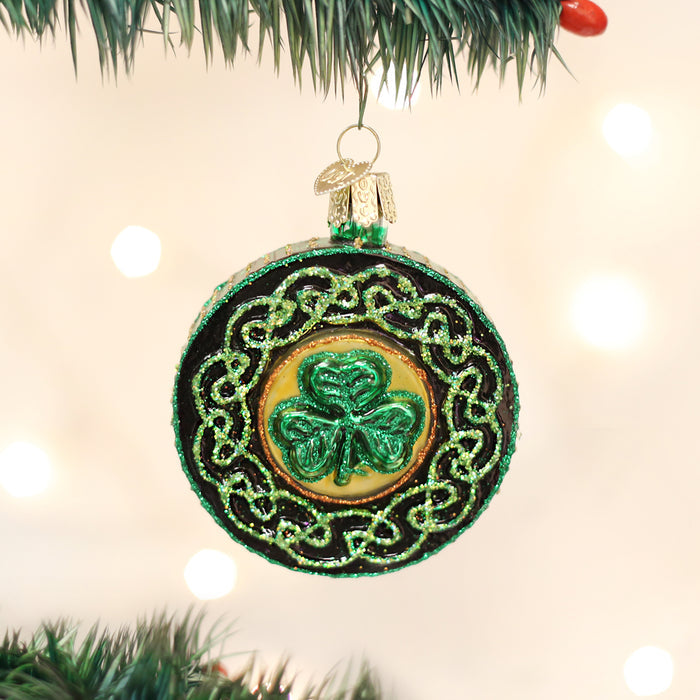 A round Christmas ornament with a green shamrock design and glittery gold and green Celtic knots hangs from a pine tree branch. The background is softly lit with white lights.