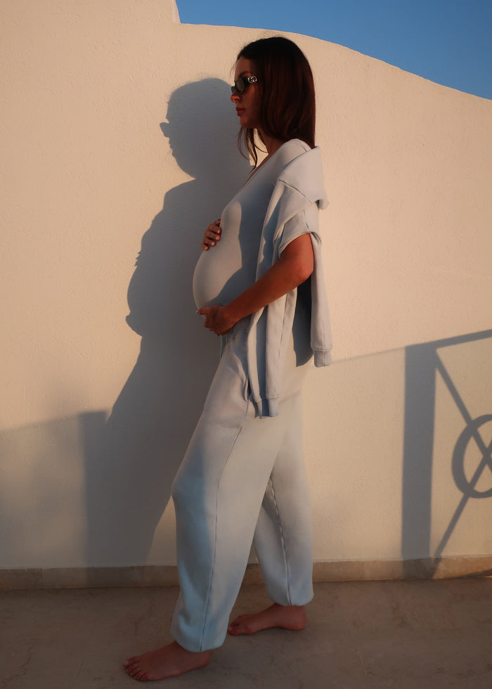 Pregnant woman in a bumpsuit light blue cotton Terry tracksuit standing against a white wall with a blue sky.