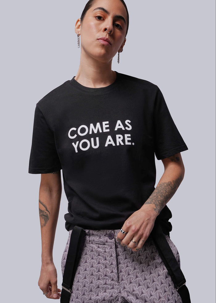 S=Person wearing a black t-shirt with 'COME AS YOU ARE.' text on a plain background