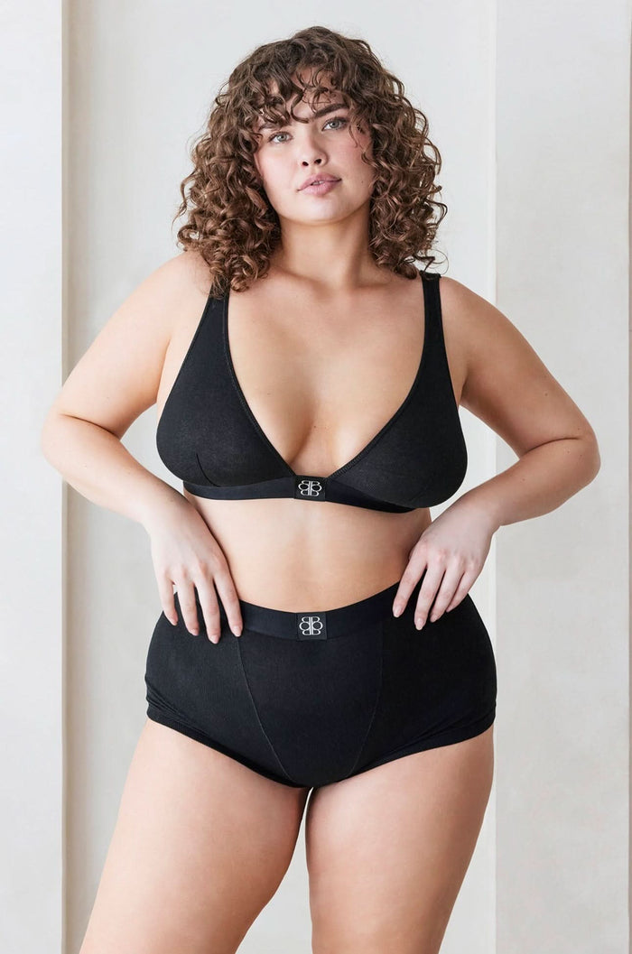 A model wearing bumpsuit maternity cotton rib plunge bralette and boy shorts in black standing against a grey background