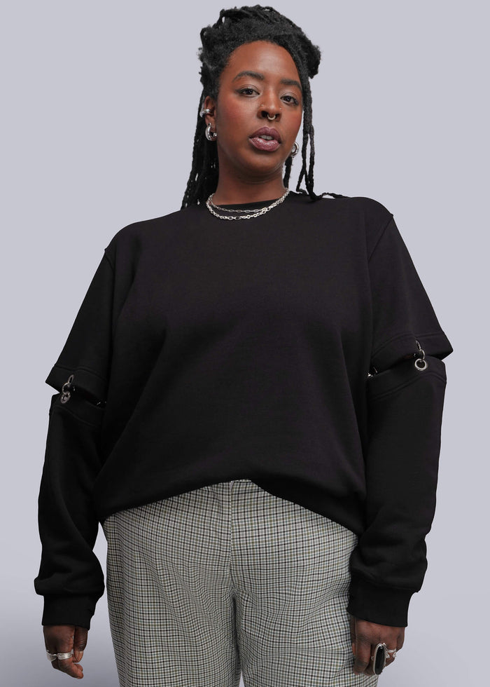 1X=A woman modeling a black crewneck sweatshirt with unique sleeve cutouts and chain details, paired with gray and white checkered trousers, against a solid gray studio background.