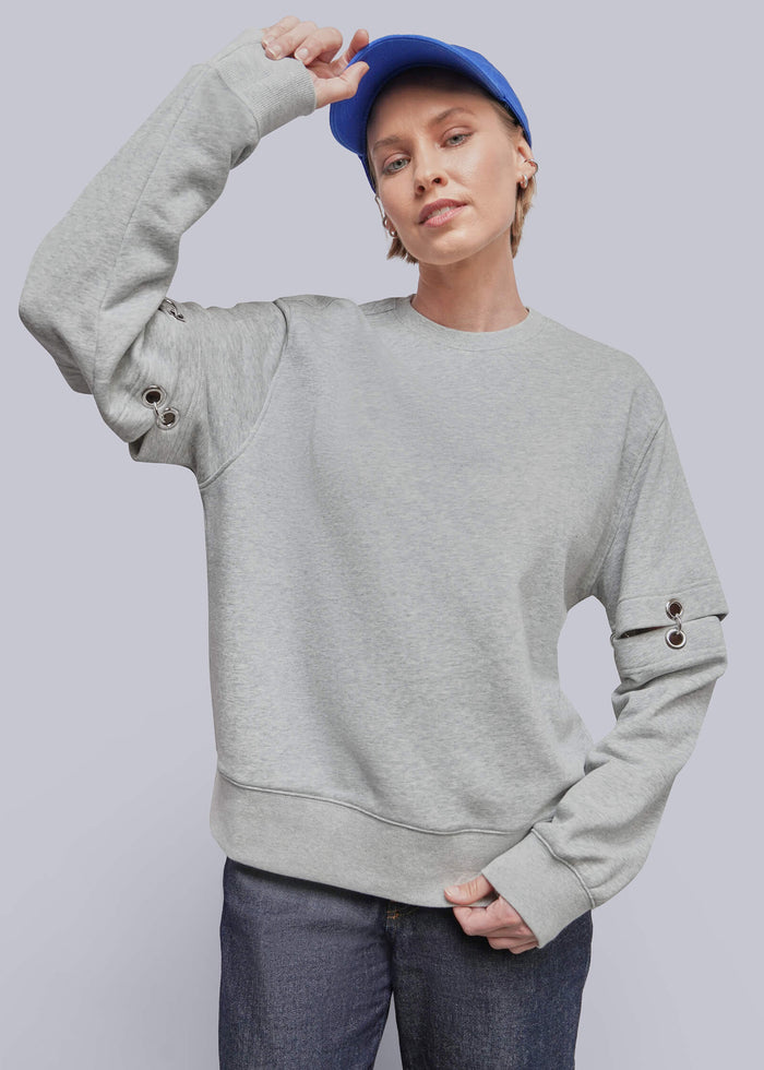 S=A model wearing a light gray crewneck sweatshirt with decorative button details on the sleeves, paired with dark blue jeans and a blue baseball cap, against a solid gray studio background.