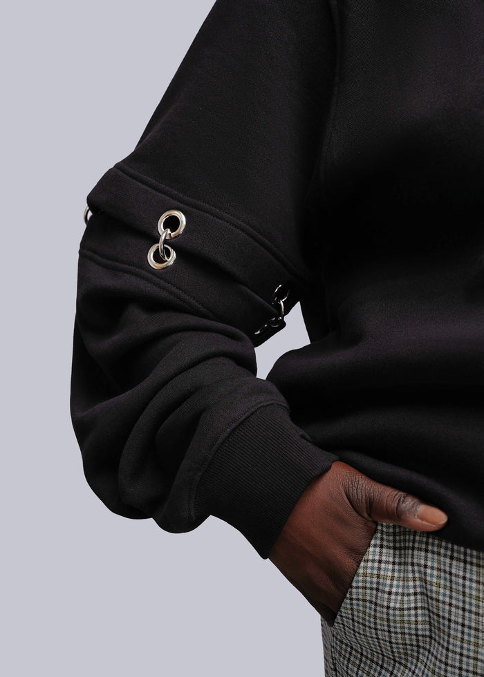 1X=Close-up of a person wearing a black long-sleeve sweatshirt with silver-tone metal grommet details on the sleeve, paired with a plaid shirt.
