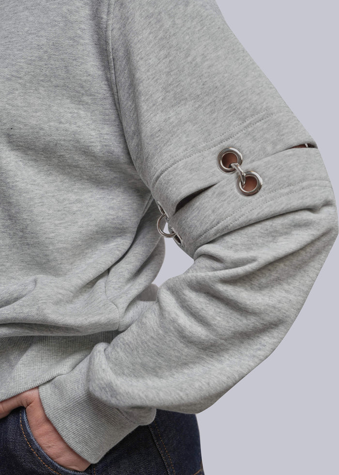 S=Close-up of a person wearing a heather gray long-sleeve sweatshirt with metal grommet details on the sleeve, paired with dark blue jeans against a light gray background.