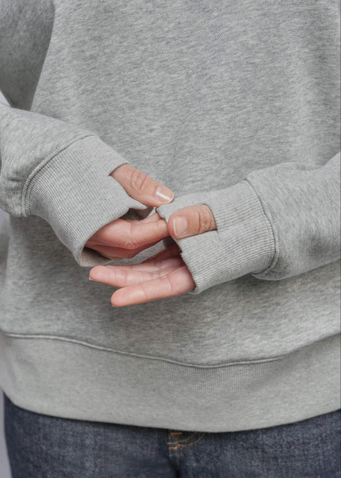 S=Close-up of a person wearing a light gray heather sweatshirt with thumbhole sleeves and dark blue jeans, showing the ribbed cuffs and hands.