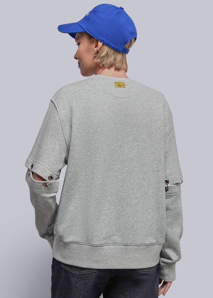 S=A person shown from the back wearing a light gray heather crewneck sweatshirt with distressed details on the sleeves, paired with dark jeans and a bright blue baseball cap, against a solid light gray studio background.