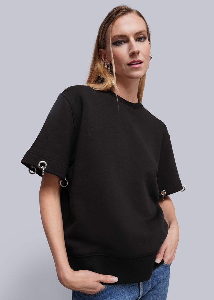 S=A woman modeling a black crewneck sweatshirt with unique sleeve details featuring silver metal rings, paired with blue jeans against a solid gray studio background.