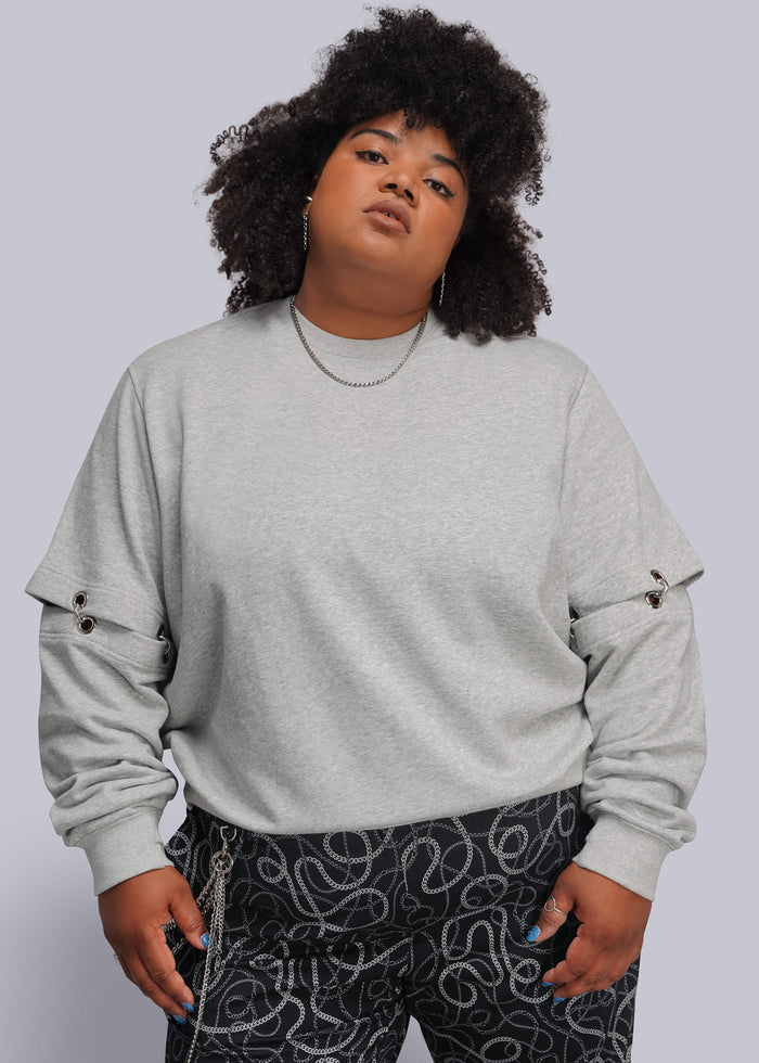 1X=A plus-size model with curly black hair wearing a light gray crewneck sweatshirt with grommet details on the sleeves and black pants with a white abstract pattern, set against a solid light gray background.