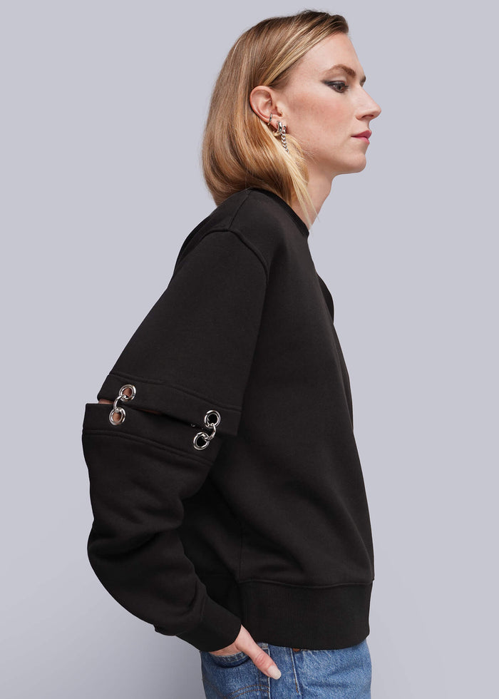 S=A woman in profile wearing a black crewneck sweatshirt with silver-tone metal ring details on the sleeve, paired with blue jeans, against a solid gray studio background.