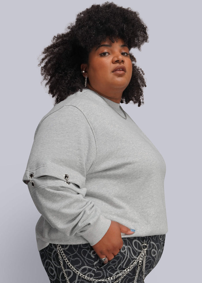 1X=A plus-size model with dark curly hair is shown from the side, wearing a light gray crewneck sweatshirt with rolled-up sleeves featuring silver metal grommet details, paired with black patterned pants with silver chain accents, against a solid light gray background.