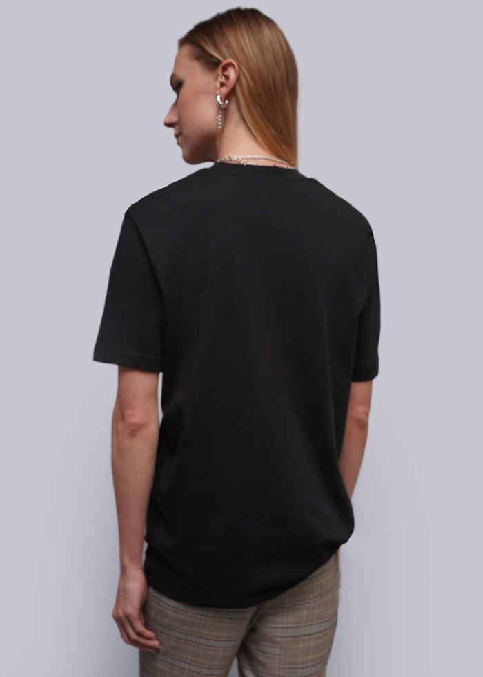 S=A model shown from the back wearing a black short-sleeve t-shirt, and beige plaid trousers, against a plain light gray studio background.