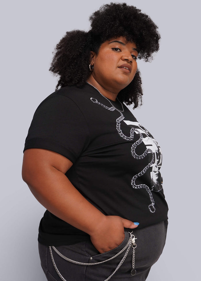 1X=A plus-size woman with dark curly hair models a black short-sleeve graphic t-shirt featuring a chain print, paired with dark gray pants and a silver wallet chain, against a solid light gray background.