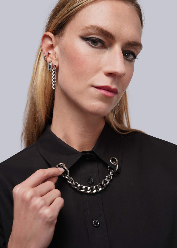 S=A close-up of a woman wearing a black button up shirt with a grommet and chain embellished collar and silver earrings.