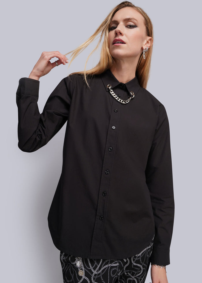 S=A woman modeling a black long-sleeve button-up shirt with a grommet and chain embellished collar, paired with black and white patterned pants, against a solid gray studio background.