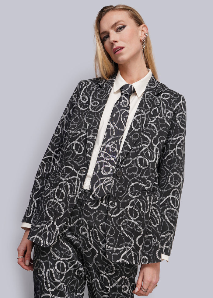 S=A woman modeling a black and white abstract patterned blazer and matching trousers, styled with a white collared shirt and tie, against a solid gray studio background.