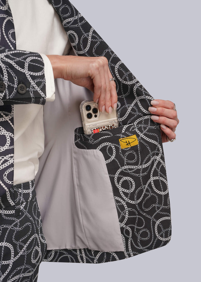 S=A close-up of a person wearing a black blazer with a white chain-link pattern, holding the lapel open to show a light gray interior pocket containing a smartphone and a small yellow tag.
