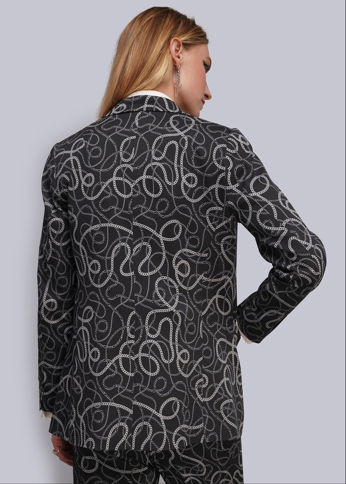S=A woman shown from the back wearing a black blazer and matching trousers with an all-over white and grey wavy chain pattern, against a light gray studio background.
